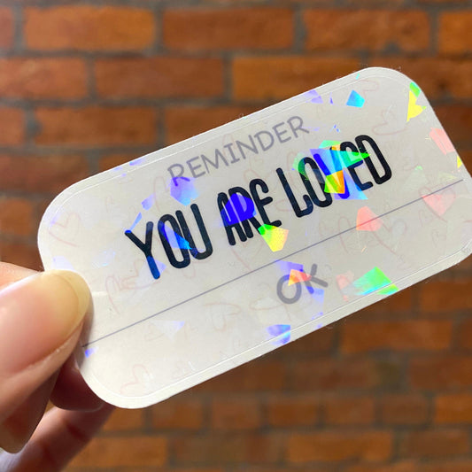 Holographic motivation sticker with the text 'YOU ARE LOVED'. Angle view
