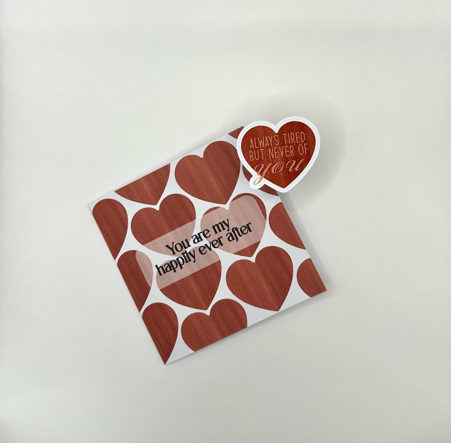 A Valentine's Day card with a heart pattern and the text 'You are my happily ever after' printed on the front. The card is white with red hearts and a visible envelope slot. Together with a heart shaped sticker with text 'always tired but never of you'