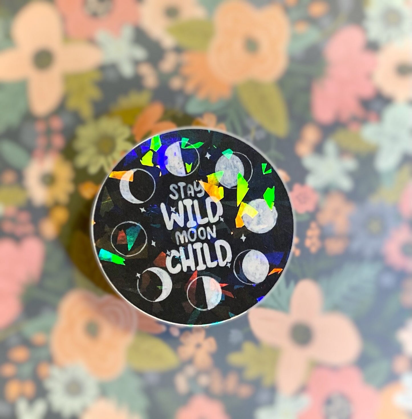 Black round celestial sticker with holographic finish, moon phaes and text 'stay wild moon child'. Frontal