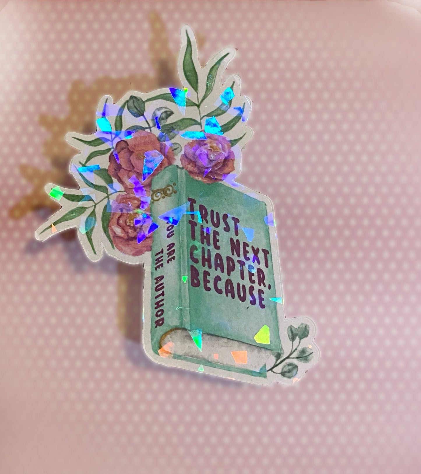 A holographic sticker with a book and floral design and the text 'TRUST THE NEXT CHAPTER BECAUSE YOU ARE THE AUTHOR' on it. Side view
