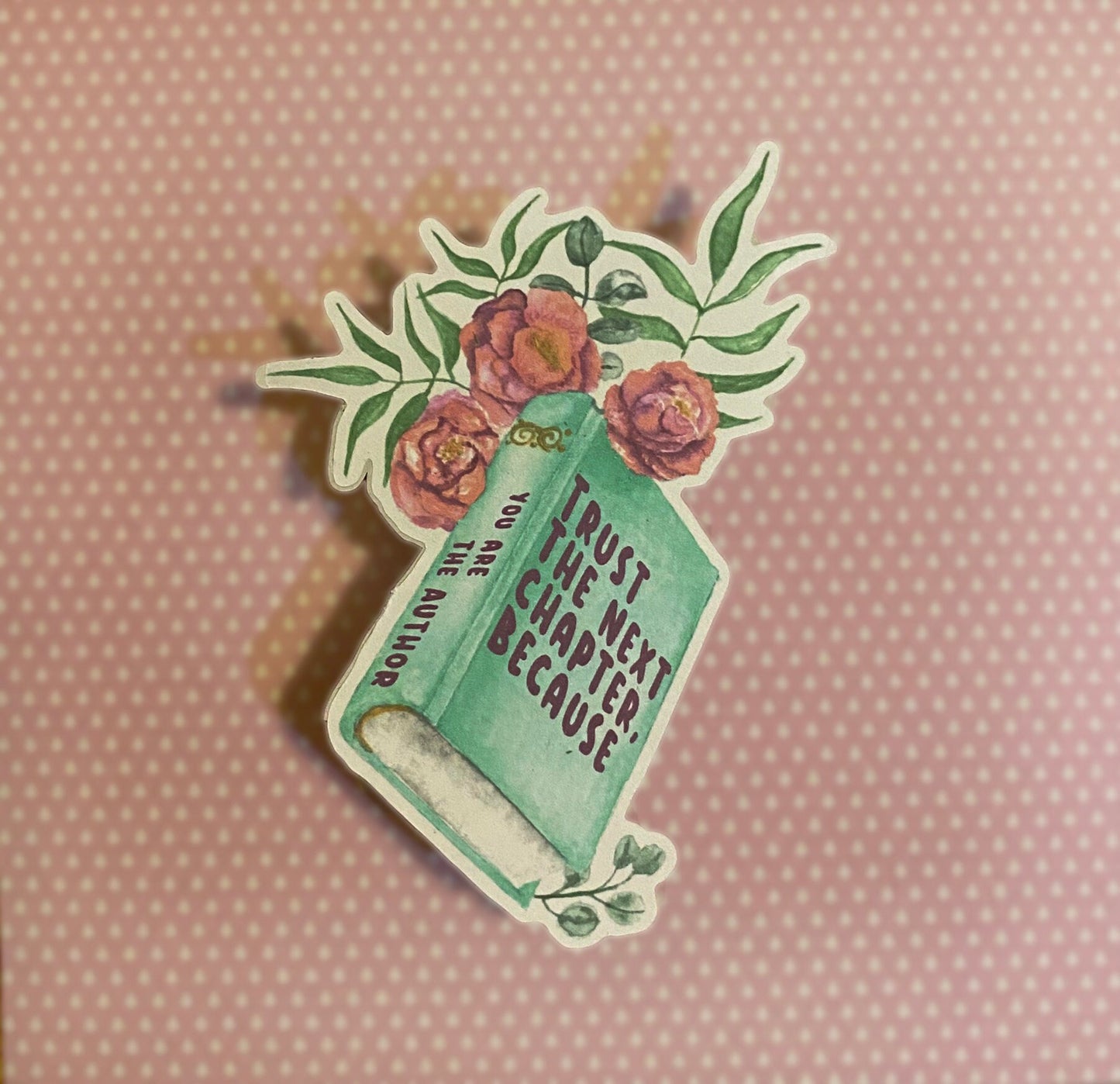 A sticker with a book and floral design and the text 'TRUST THE NEXT CHAPTER BECAUSE YOU ARE THE AUTHOR' on it, against a brick wall background. Side view