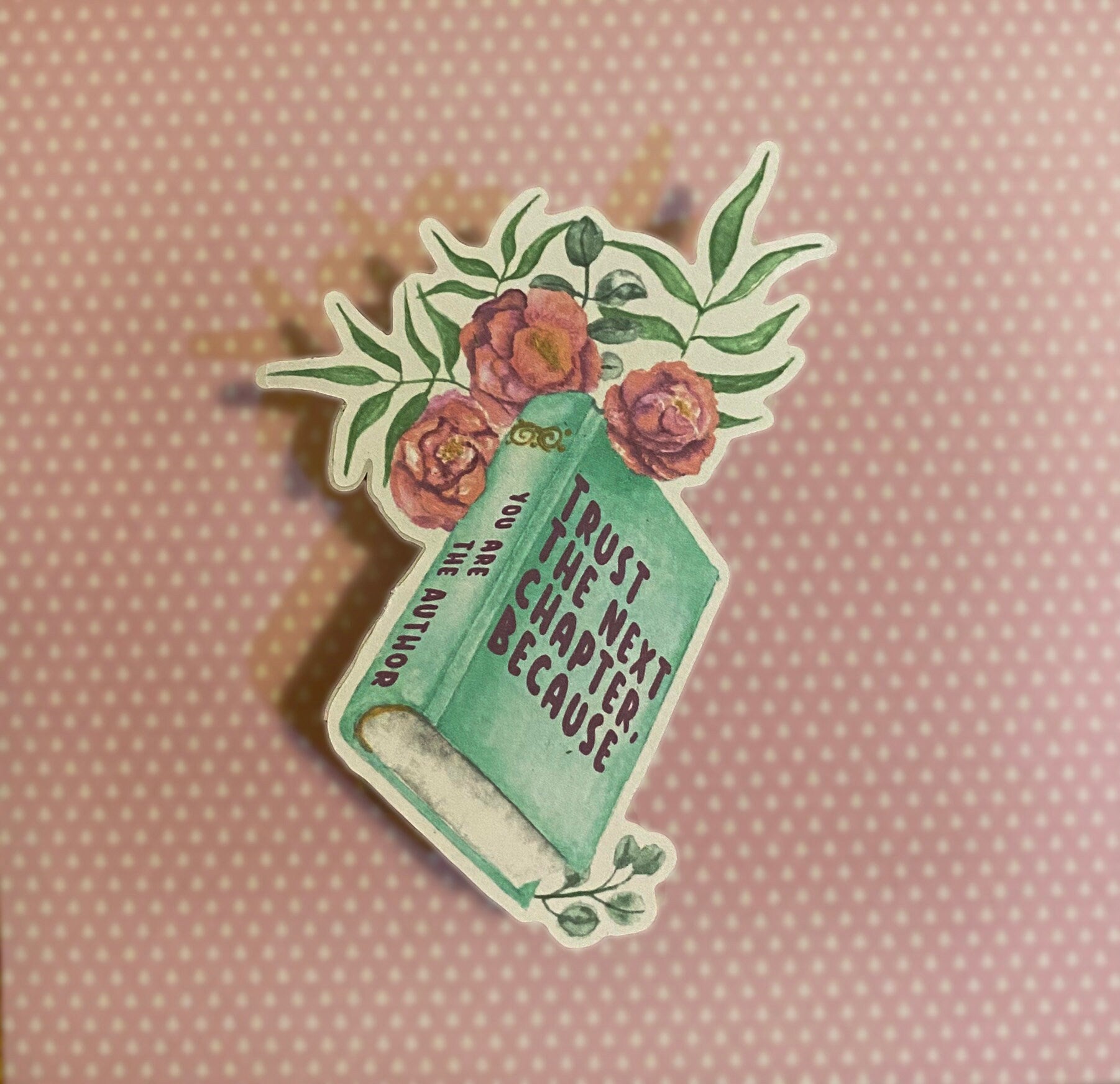 A sticker with a book and floral design and the text 'TRUST THE NEXT CHAPTER BECAUSE YOU ARE THE AUTHOR' on it, against a brick wall background. Side view