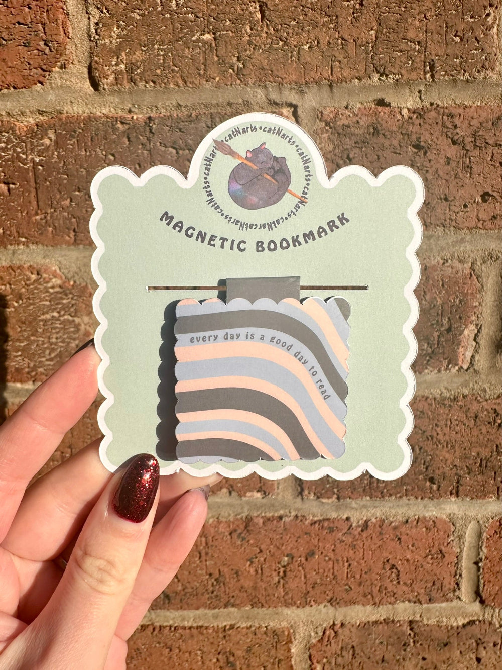 Hand holding a colorful magnetic bookmark with a text 'every day is a good day to read' against a brick wall background
