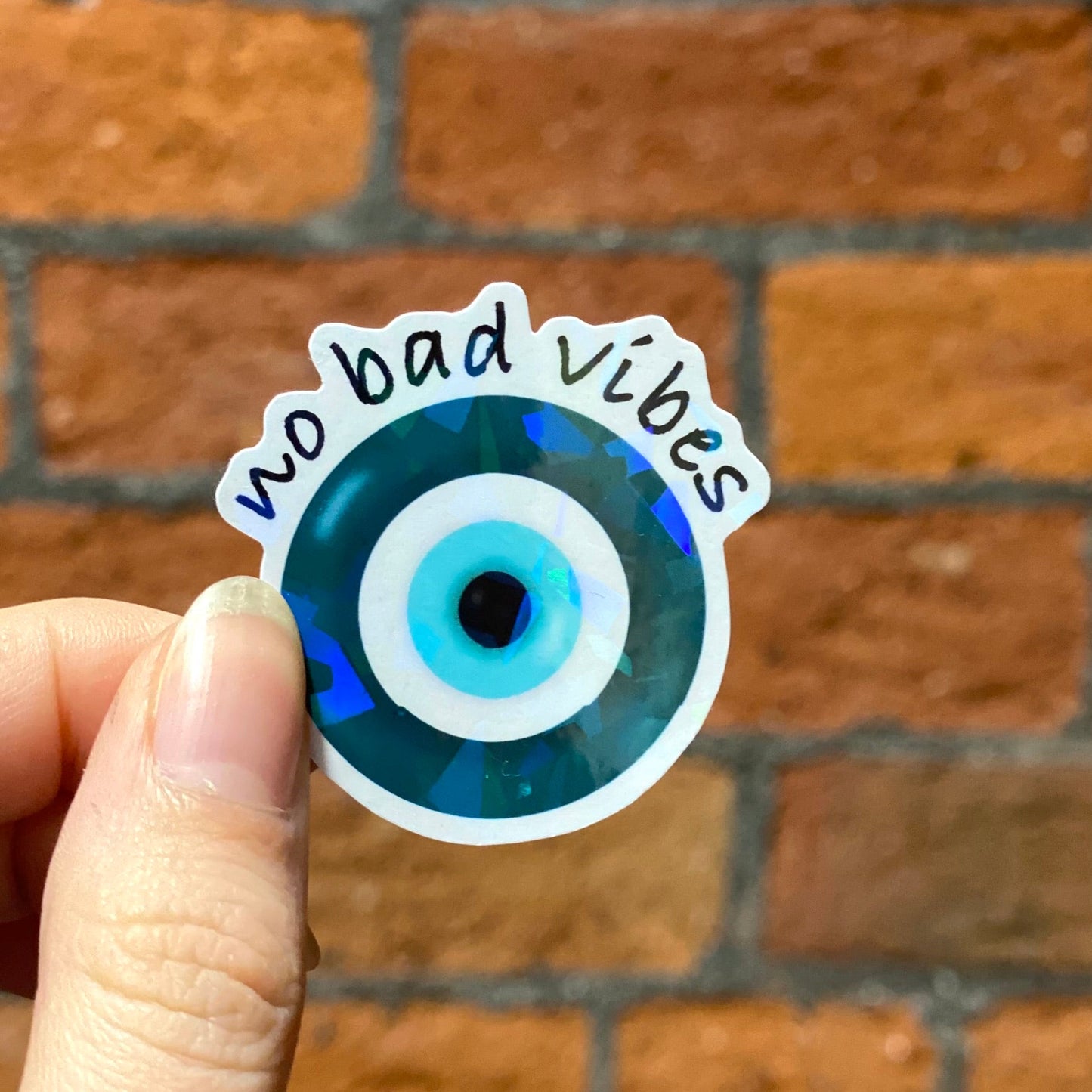 Aesthetic round holographic sticker with a blue and black evil eye design and the text 'no bad vibes' . Frontal
