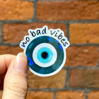 Aesthetic round holographic sticker with a blue and black evil eye design and the text 'no bad vibes' . Frontal