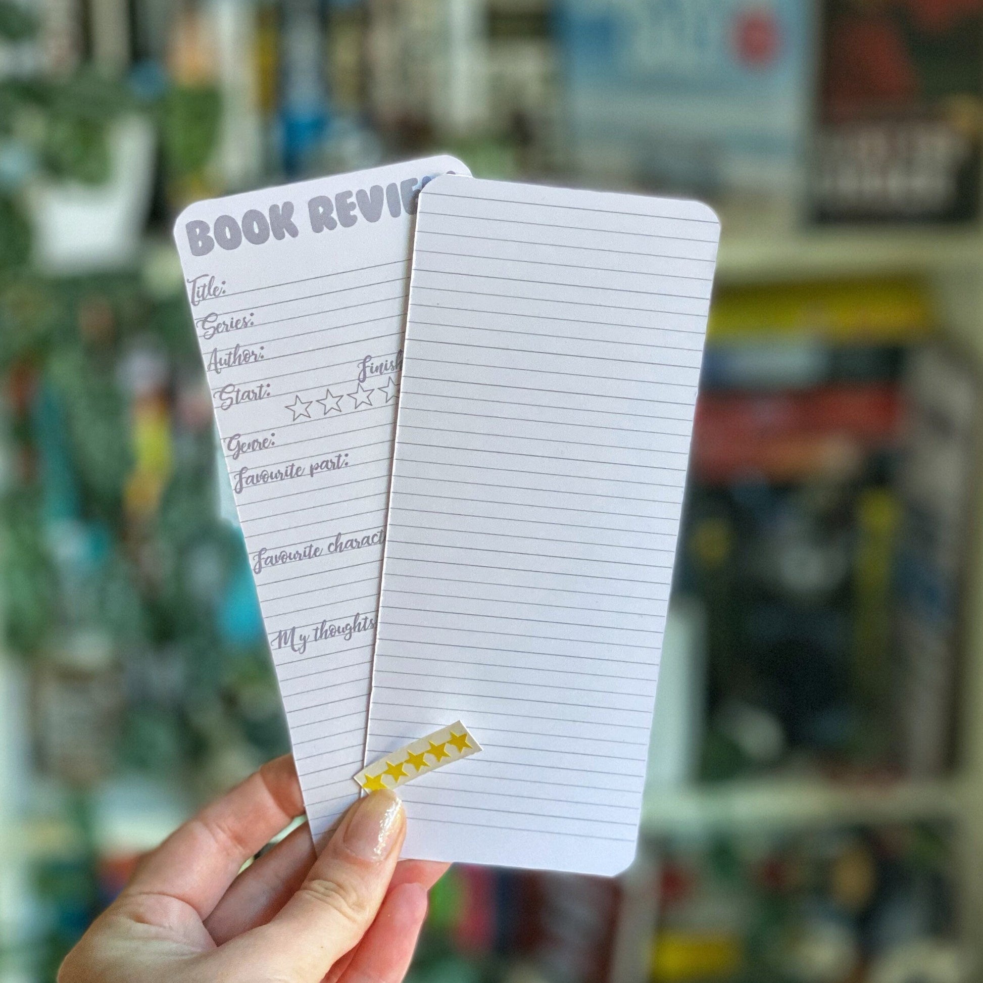 A hand holding a double-sided bookmark with lined areas for notes and a set of yellow star stickers for rating, with a blurred background of books.