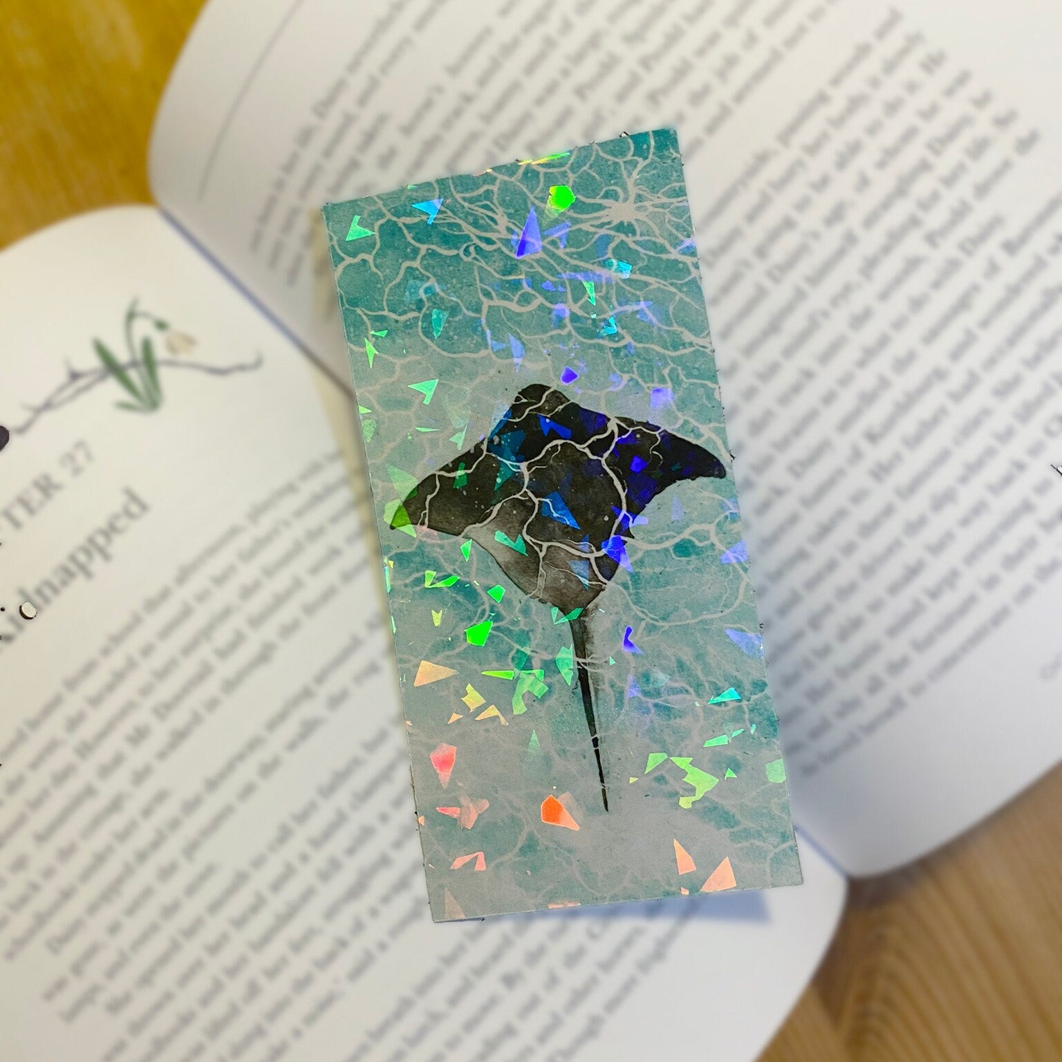 Holographic finish bookmark with a marine ray fish design, on top of a book. Top view