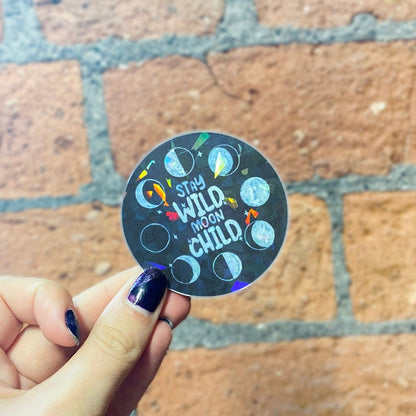 Black round celestial sticker with holographic finish, moon phaes and text 'stay wild moon child'. Frontal