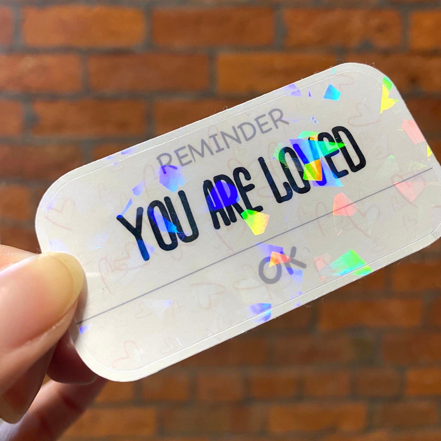 Holographic motivation sticker with the text 'YOU ARE LOVED'. Angle view