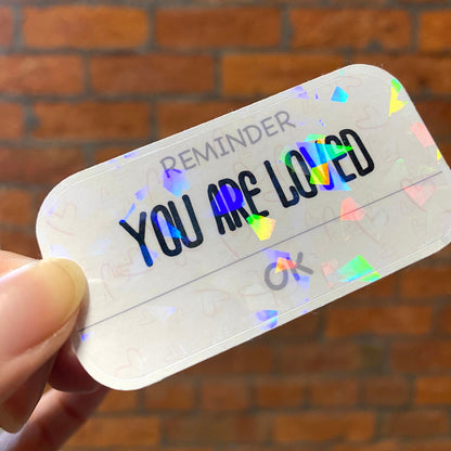 Holographic motivation sticker with the text 'YOU ARE LOVED'. Angle view