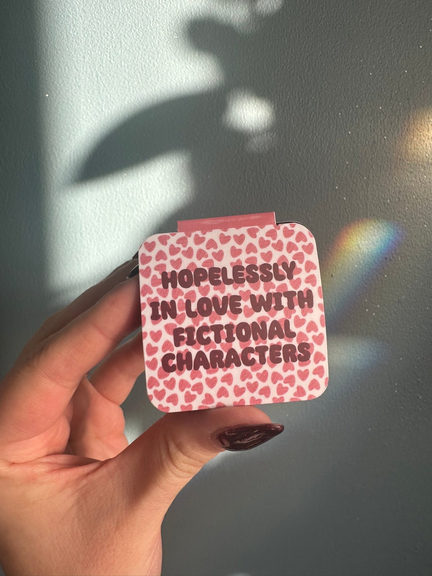 Hand holding a square magnetic bookmark with heart patterns and 'Hopelessly in love with fictional characters' text against a light background