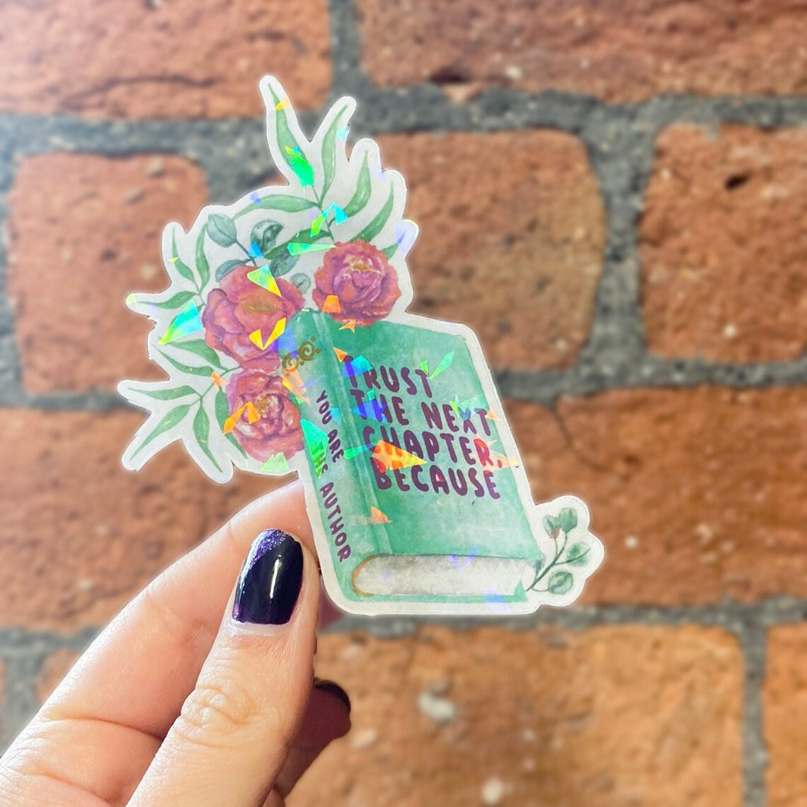 A holographic sticker with a book and floral design and the text 'TRUST THE NEXT CHAPTER BECAUSE YOU ARE THE AUTHOR' on it. Frontal