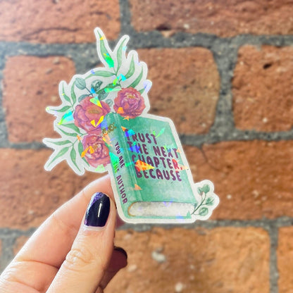 A holographic sticker with a book and floral design and the text 'TRUST THE NEXT CHAPTER BECAUSE YOU ARE THE AUTHOR' on it. Frontal