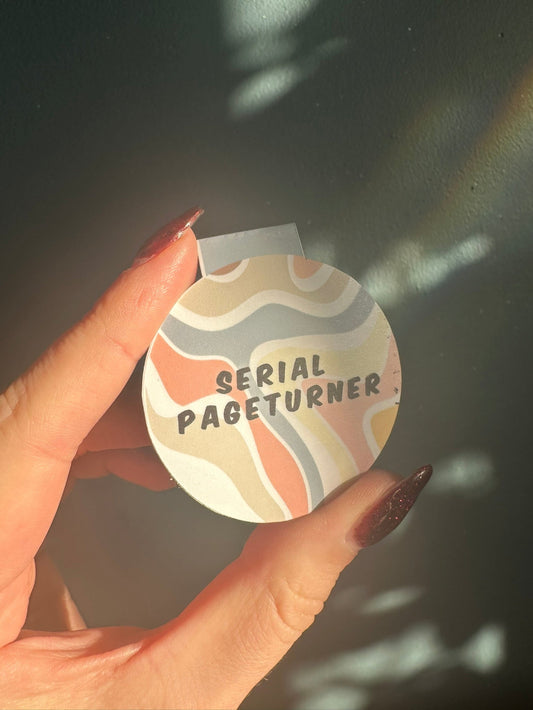 Hand holding a round magnetic bookmark with 'Serial Pageturner' text against a dark background