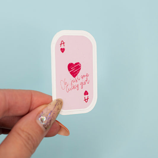  a small die-cut sticker featuring a pink playing card design with a red heart and the phrase 'Oh, just one lucky girl' against a light blue background.