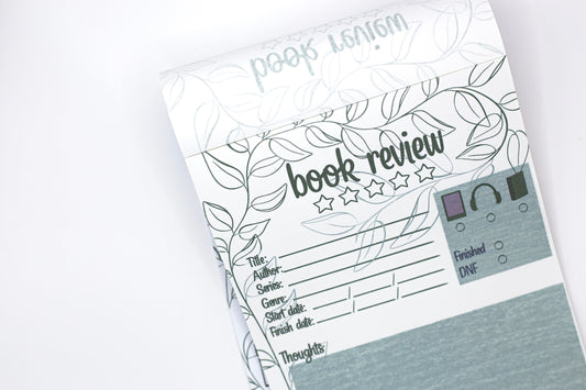 A close-up of a book review planner pad with printed fields for title, author, series, genre, start and end dates, and a small checklist box, shown on a white background.
