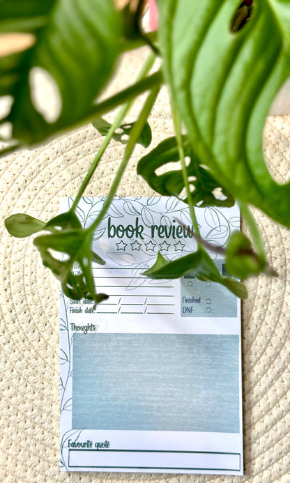 Notepad with book review template under a leafy plant on a textured surface