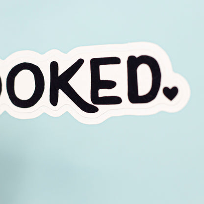 A die-cut sticker with the word 'booked.' in black handwritten-style lettering on a white background, shown against a soft light blue backdrop.