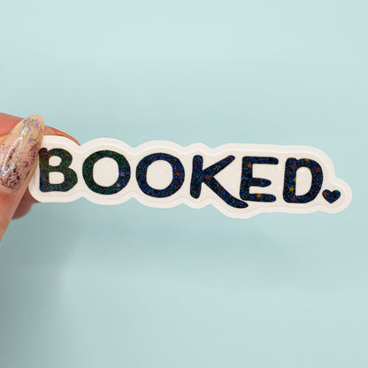 a die-cut sticker that reads 'BOOKED.' in black letters with a holographic overlay with a white outline, set against a solid light blue background.