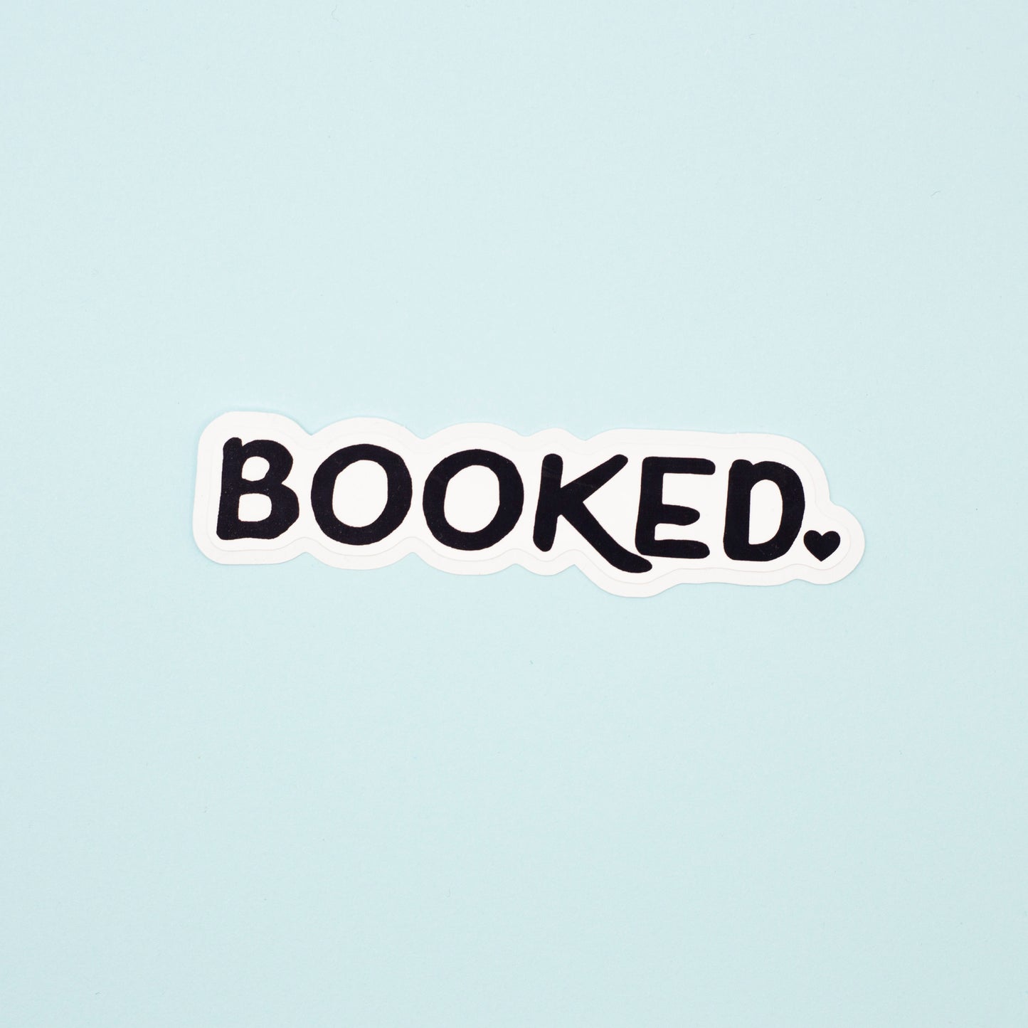 A die-cut sticker with the word 'BOOKED.' in black handwritten-style lettering on a white background, centered on a pale blue backdrop.