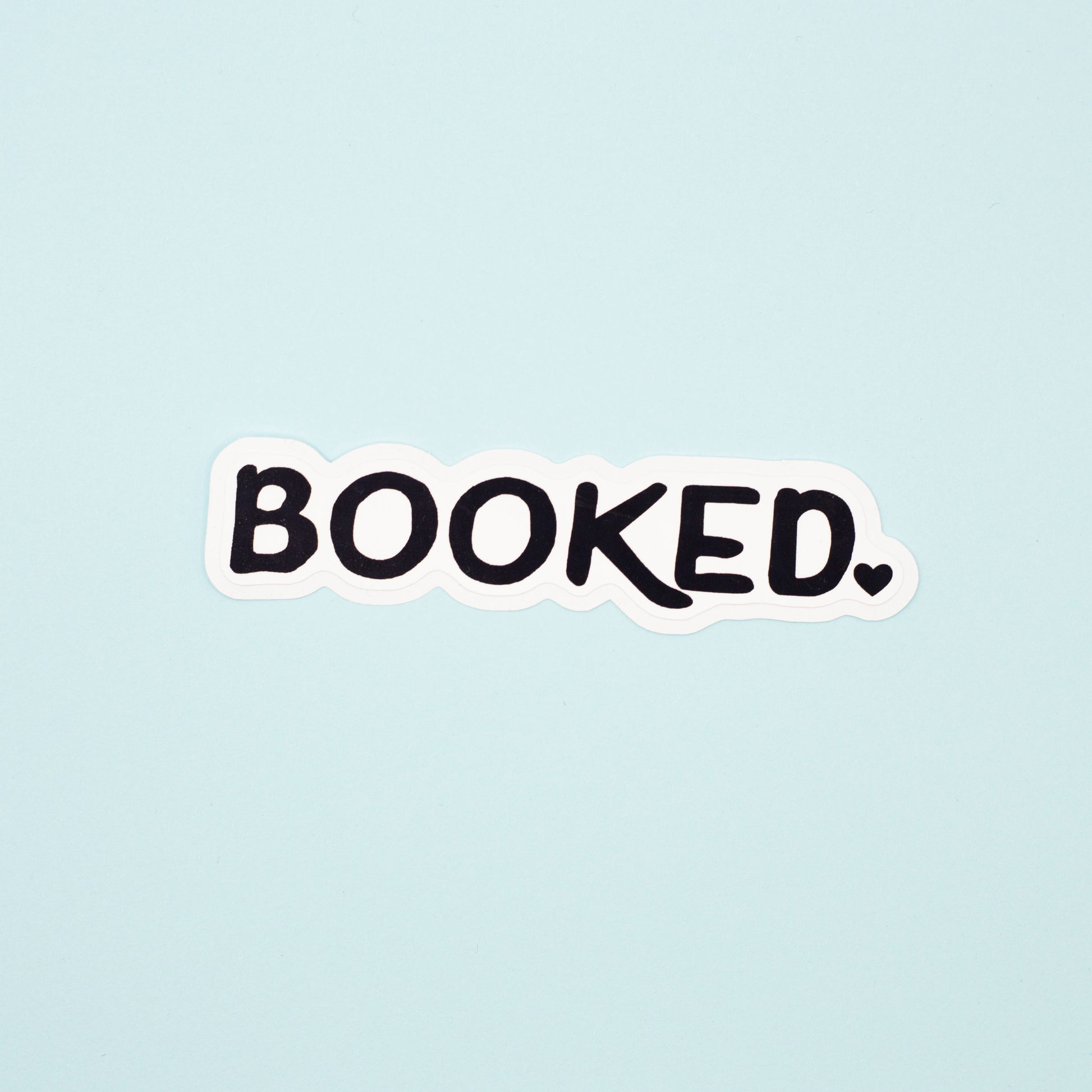 A die-cut sticker with the word 'BOOKED.' in black handwritten-style lettering on a white background, centered on a pale blue backdrop.