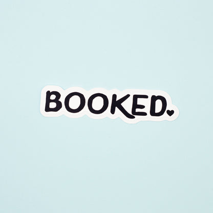 A die-cut sticker with the word 'BOOKED.' in black handwritten-style lettering on a white background, centered on a pale blue backdrop.