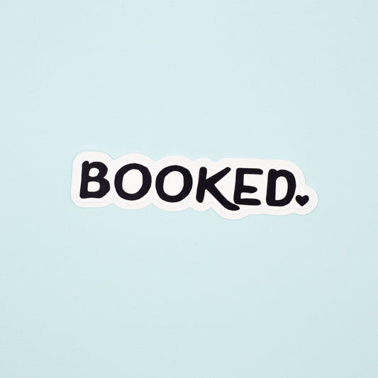 A die-cut sticker with the word 'BOOKED.' in black handwritten-style lettering on a white background, centered on a pale blue backdrop.