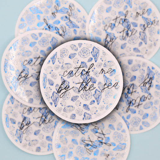 round sticker decorated with metalic blue coastal patterns and the cursive text 'catch me by the sea' in black, arranged in a circular flat-lay pattern on a light blue background.