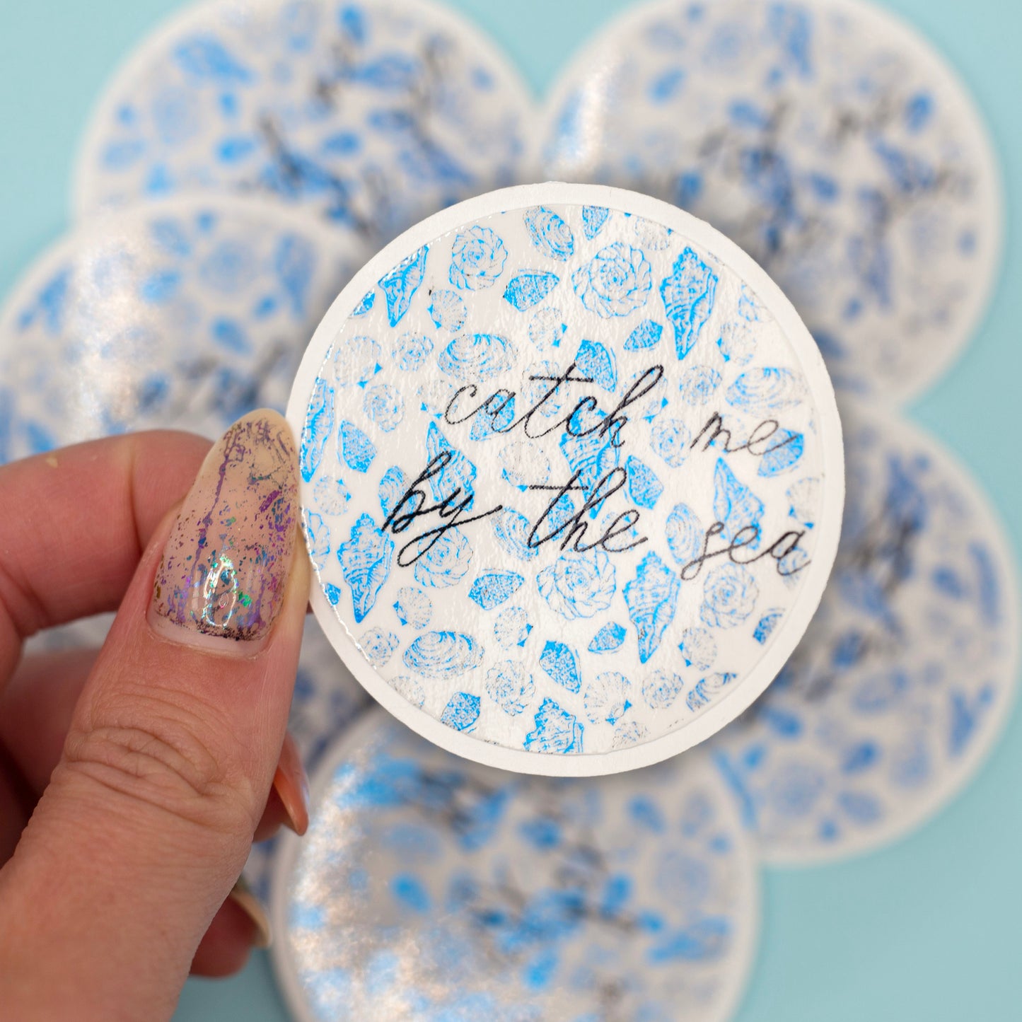  a round sticker that reads 'catch me by the sea' in black script over a metalic blue seashell pattern. More round stickers are blurred in the background against a light blue backdrop.