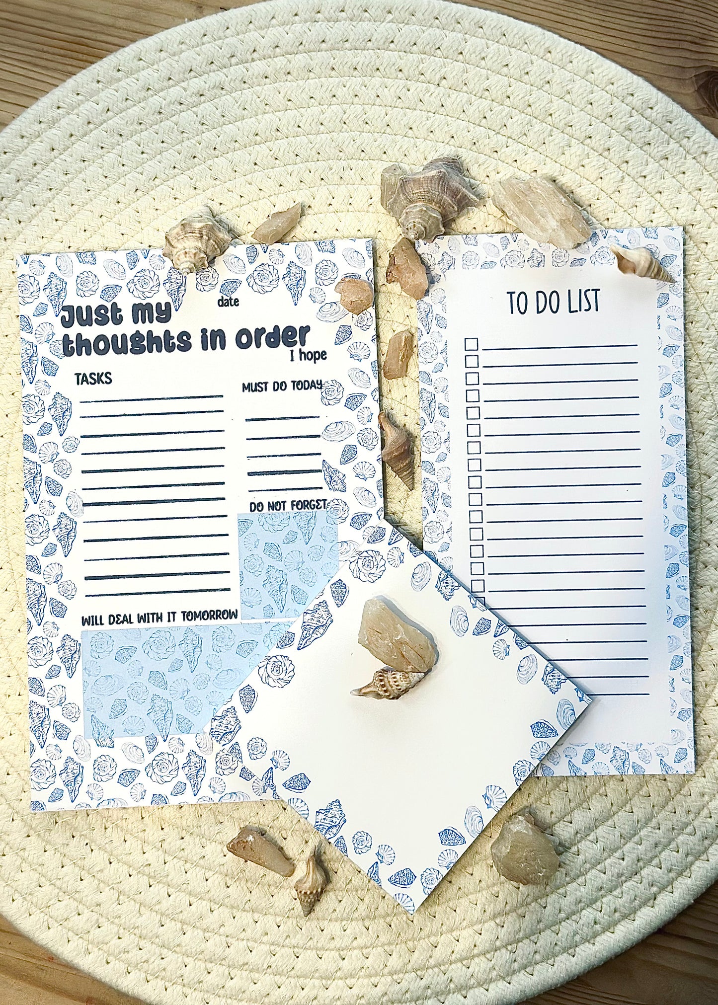 three note pads with coastal design. One square, one A5 with just my thoughts in order text and one slim pad with to-do list on a woven surface with seashells.