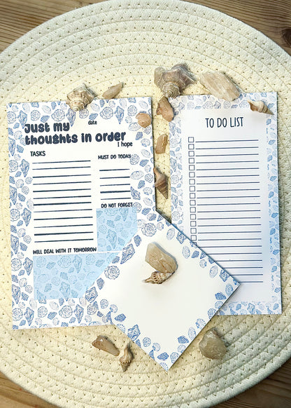 three note pads with coastal design. One square, one A5 with just my thoughts in order text and one slim pad with to-do list on a woven surface with seashells.