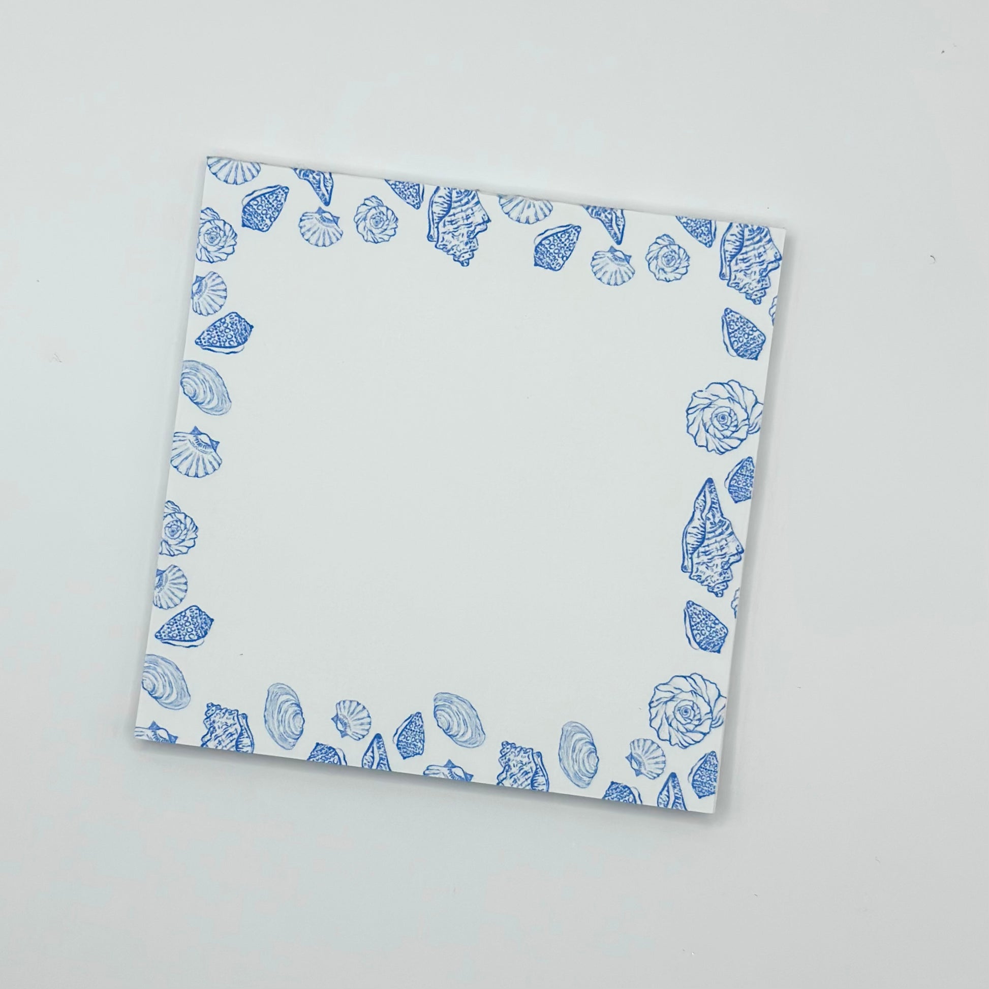 A square memo pad with a blue seashell border pattern, photographed from an angle on a light gray background.