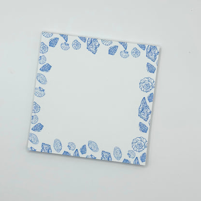 A square memo pad with a blue seashell border pattern, photographed from an angle on a light gray background.