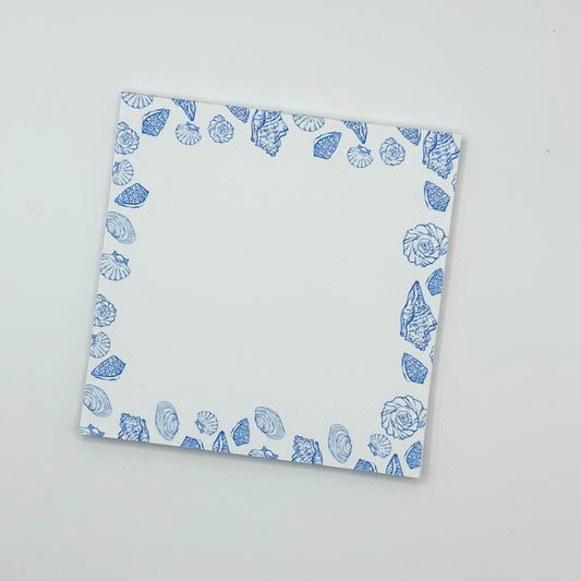 A square memo pad with a blue seashell border pattern, photographed from an angle on a light gray background.