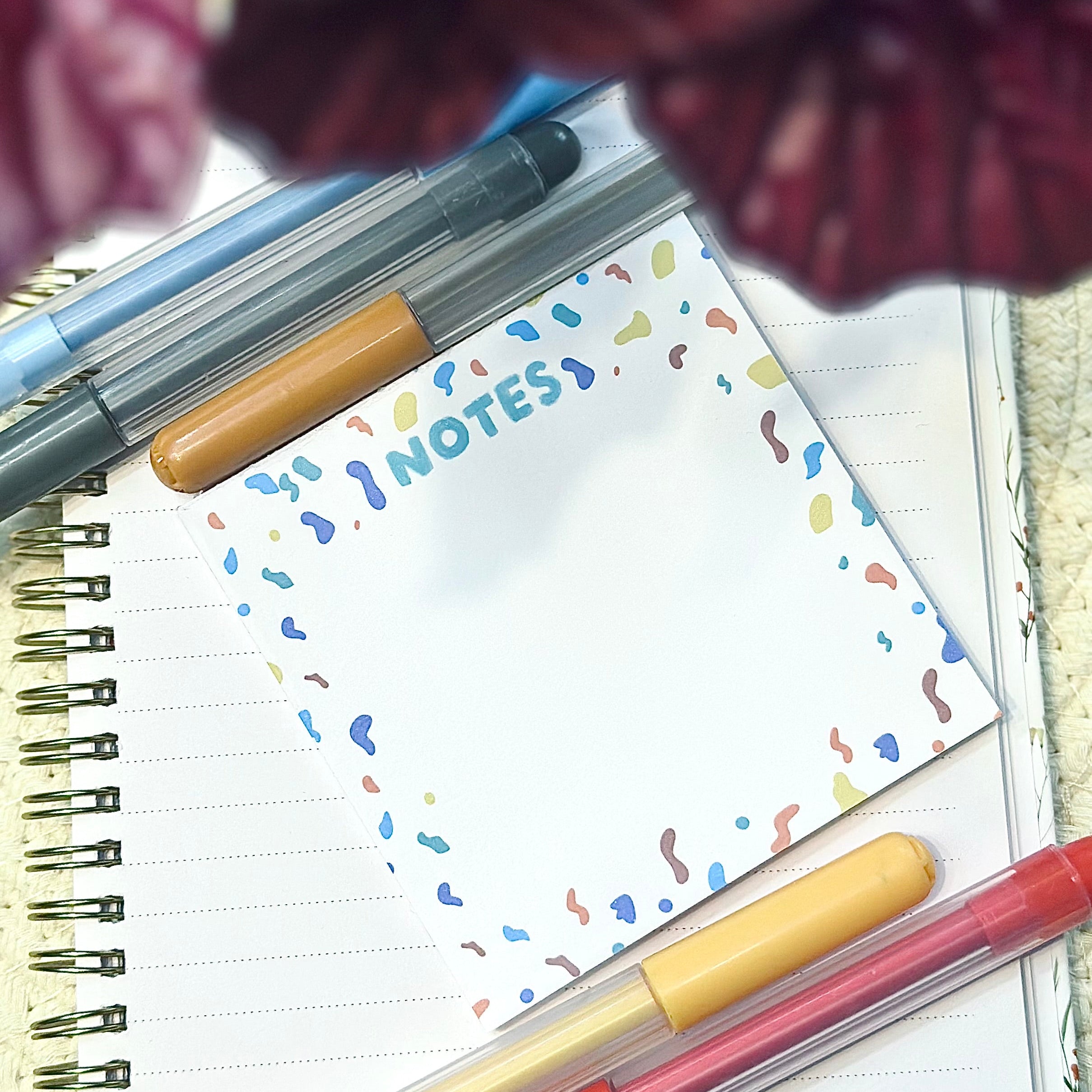 Notebook with pens and a colorful 'NOTES' confetti themed square memo pad on a textured surface.