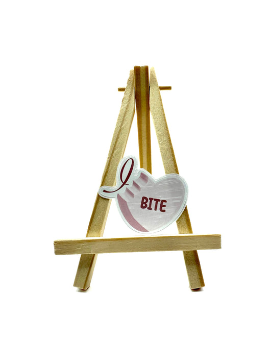 Heart-shaped sticker with 'I Bite' text on a wooden stand against a white background