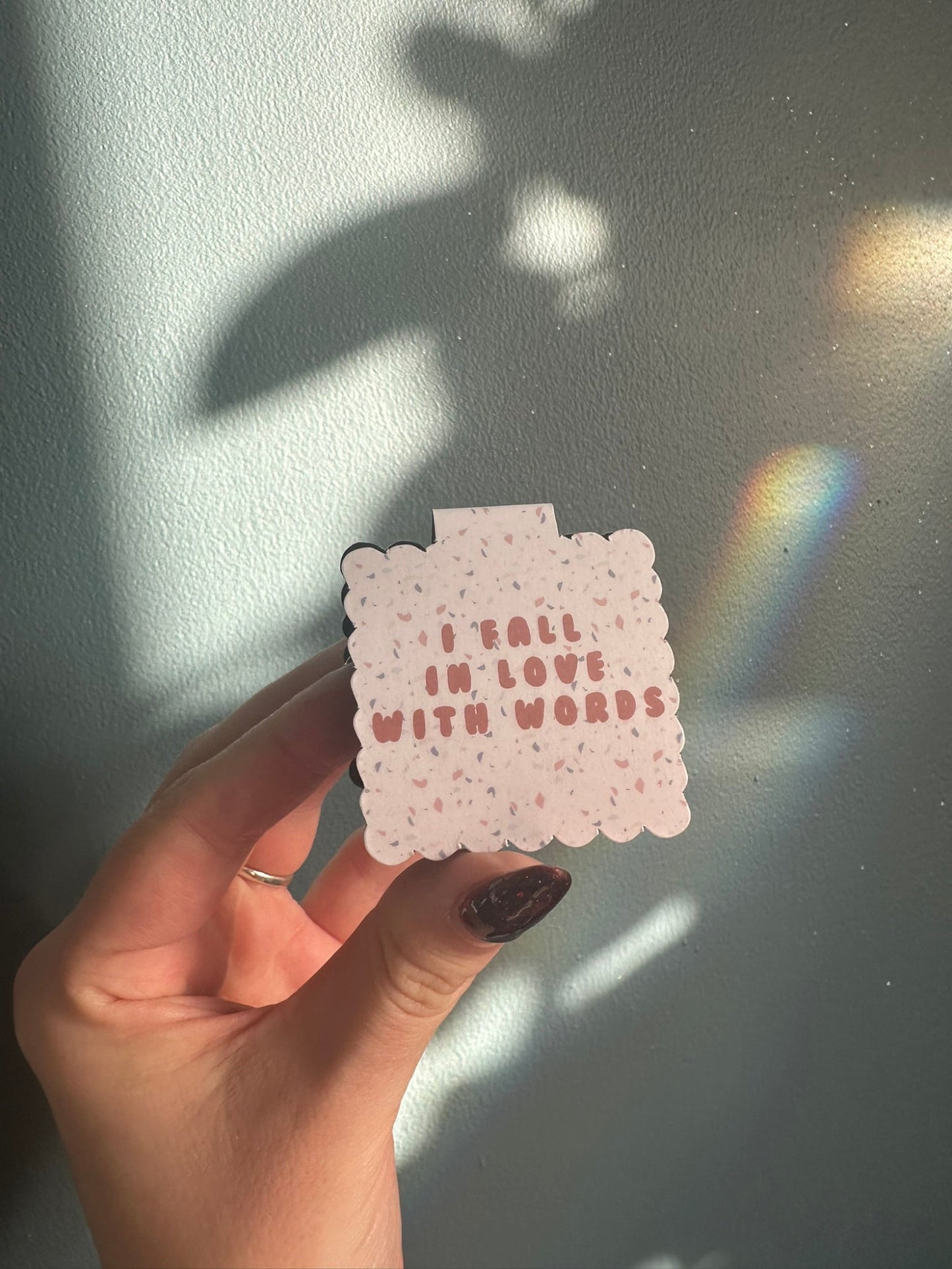 Hand holding a small terrazzo style magnetic bookmark with 'I fall in love with words' against a plain blue background