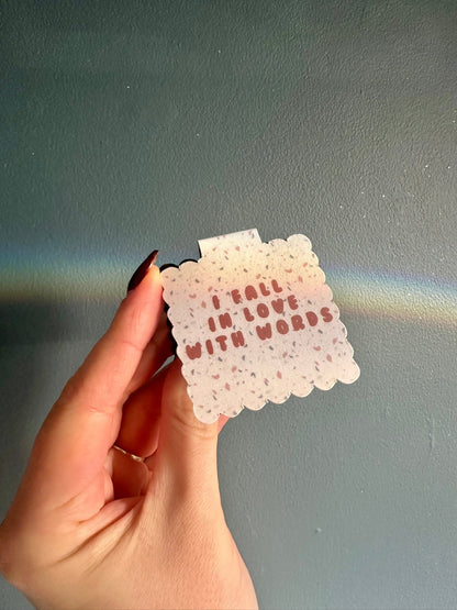 Hand holding a small terrazzo style magnetic bookmark with 'I fall in love with words' against a plain blue background