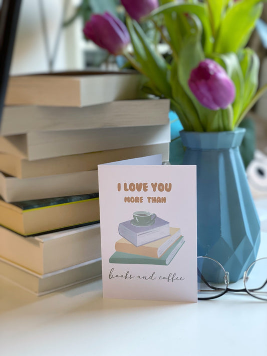 A greeting card with the text 'I LOVE YOU MORE THAN books and coffee' displayed in front of a stack of books and a vase with purple flowers.