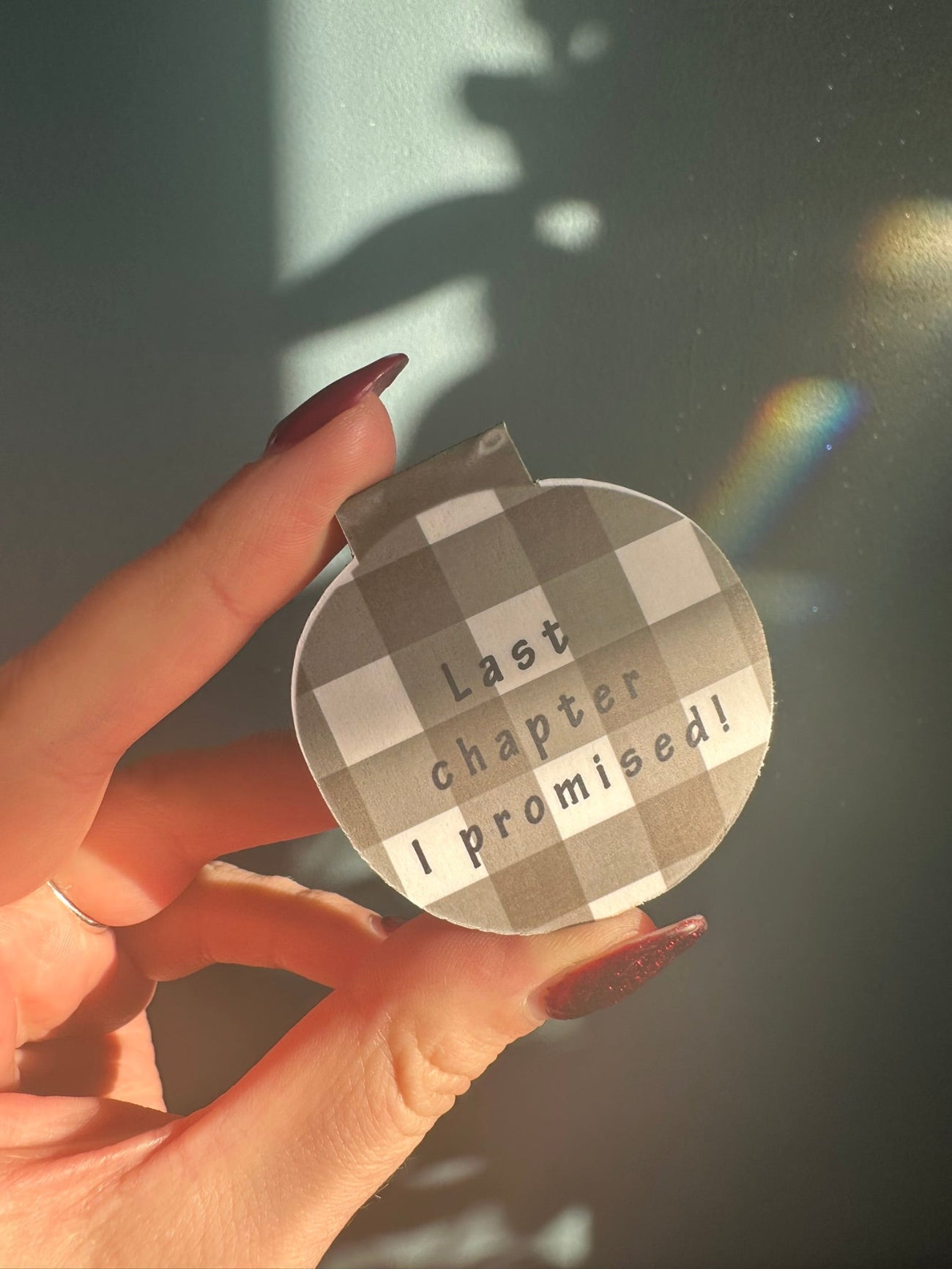 Hand holding a round magnetic bookmark with text 'last chapter I promised'  against a blurred background