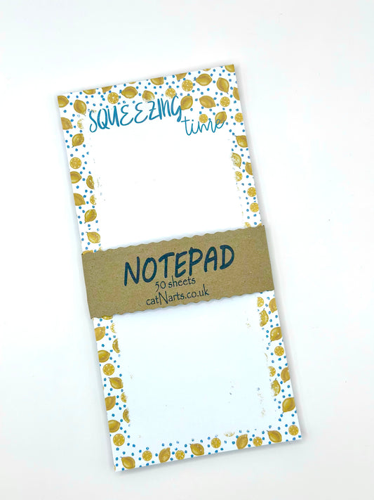 Notepad with a white background, blue flowers and lemons patterned edges. The top reads 'SQUEEZING time' in blue script, and a brown paper band across the center reads 'NOTEPAD 50 sheets callNarts.