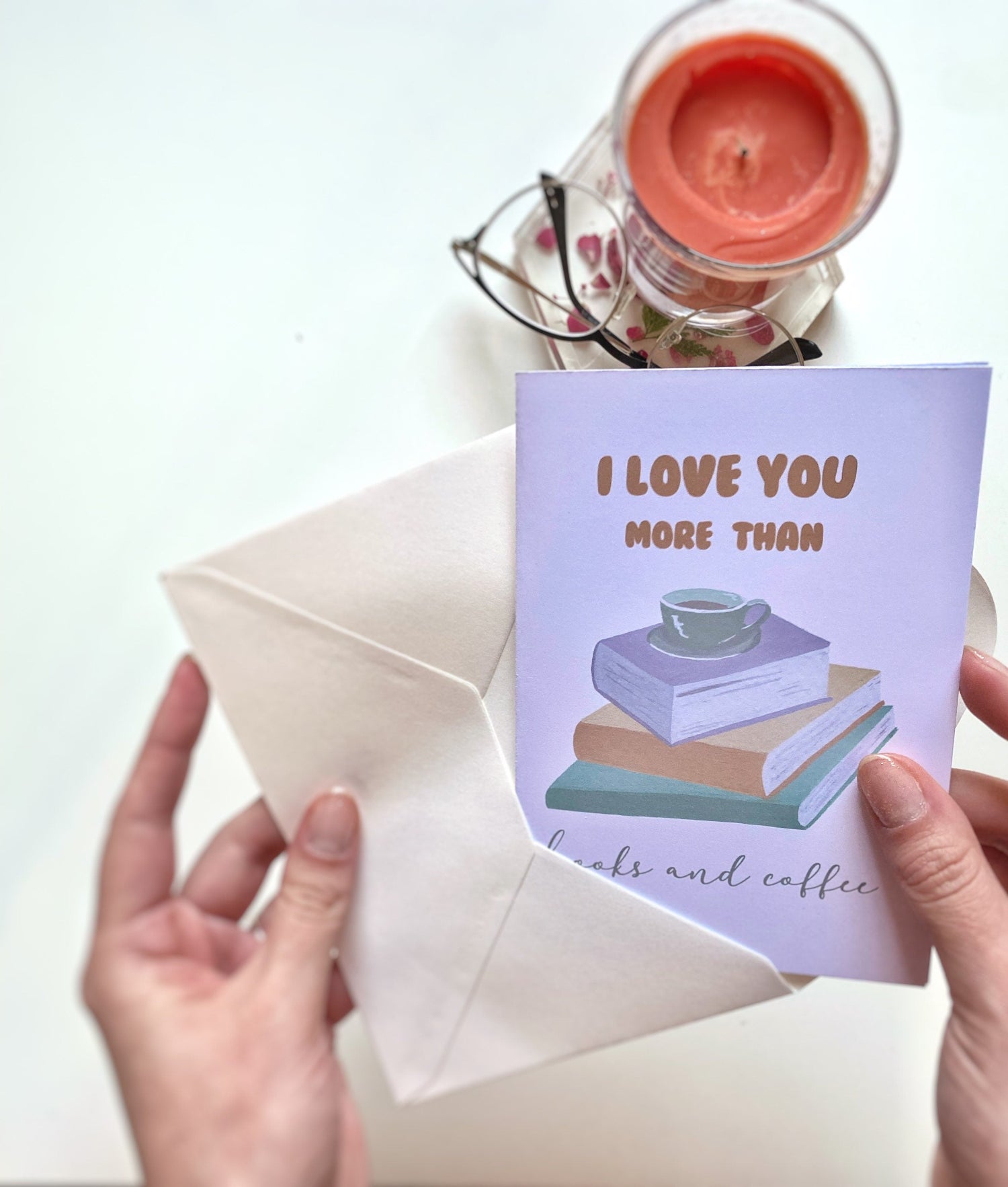 A greeting card with the text 'I LOVE YOU MORE THAN books and coffee' displayed in front of a stack of books and a vase with purple flowers.