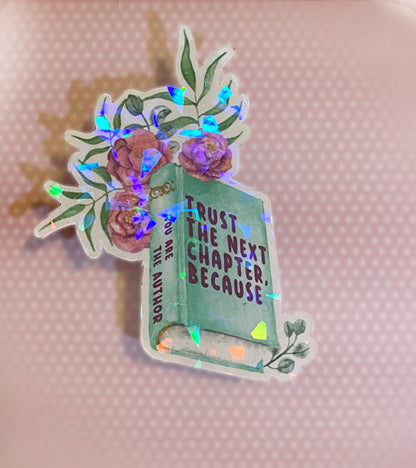 A holographic sticker with a book and floral design and the text 'TRUST THE NEXT CHAPTER BECAUSE YOU ARE THE AUTHOR' on it. Side view