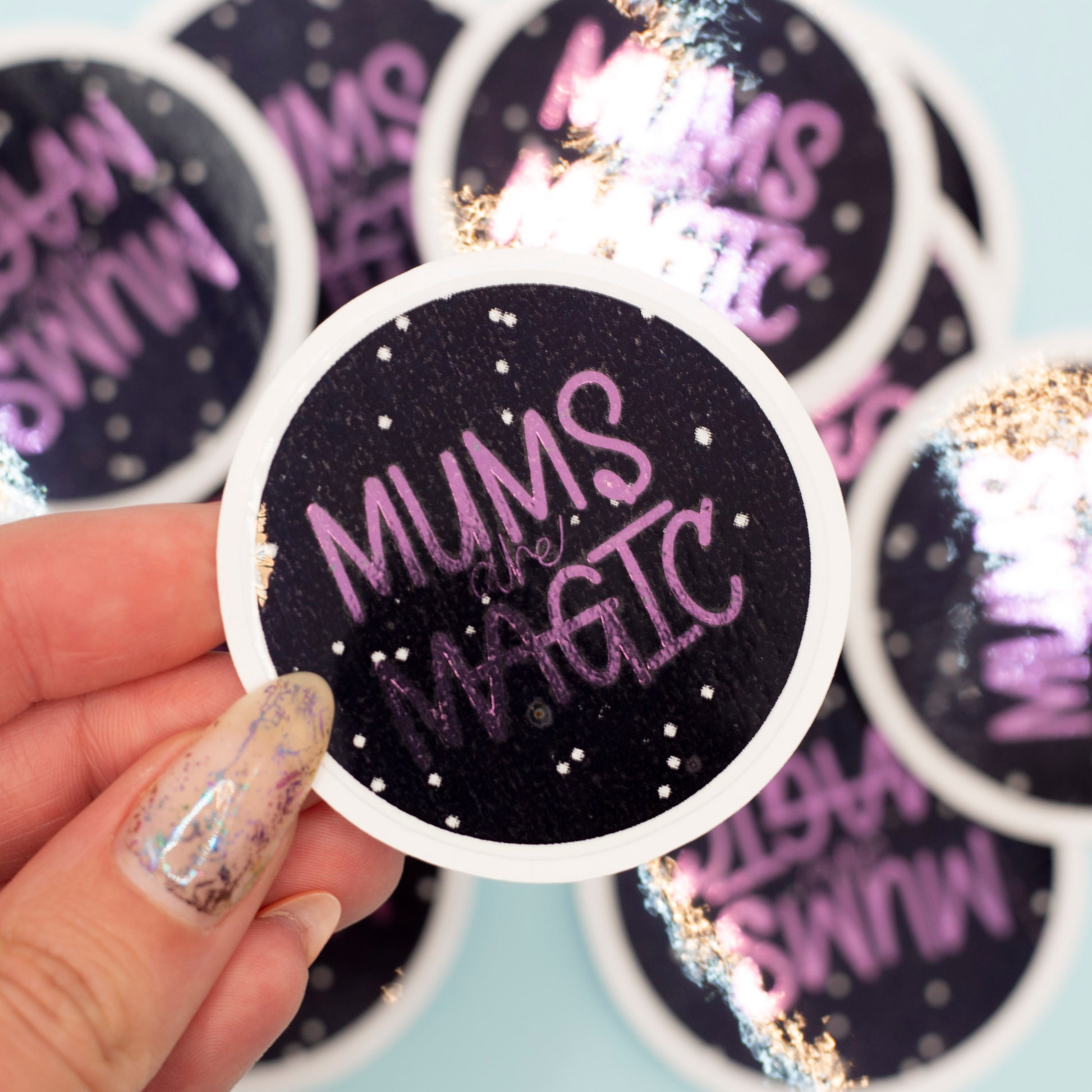 a round sticker that reads 'MUMS are MAGIC' in purple metalic text on a black background with white speckles and a white border. More identical stickers are blurred in the background against a pale blue surface.
