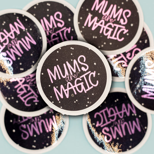 a round sticker that reads 'MUMS are MAGIC' in purple metalic text on a black background with white speckles and a white border. More identical stickers are blurred in the background against a pale blue surface.