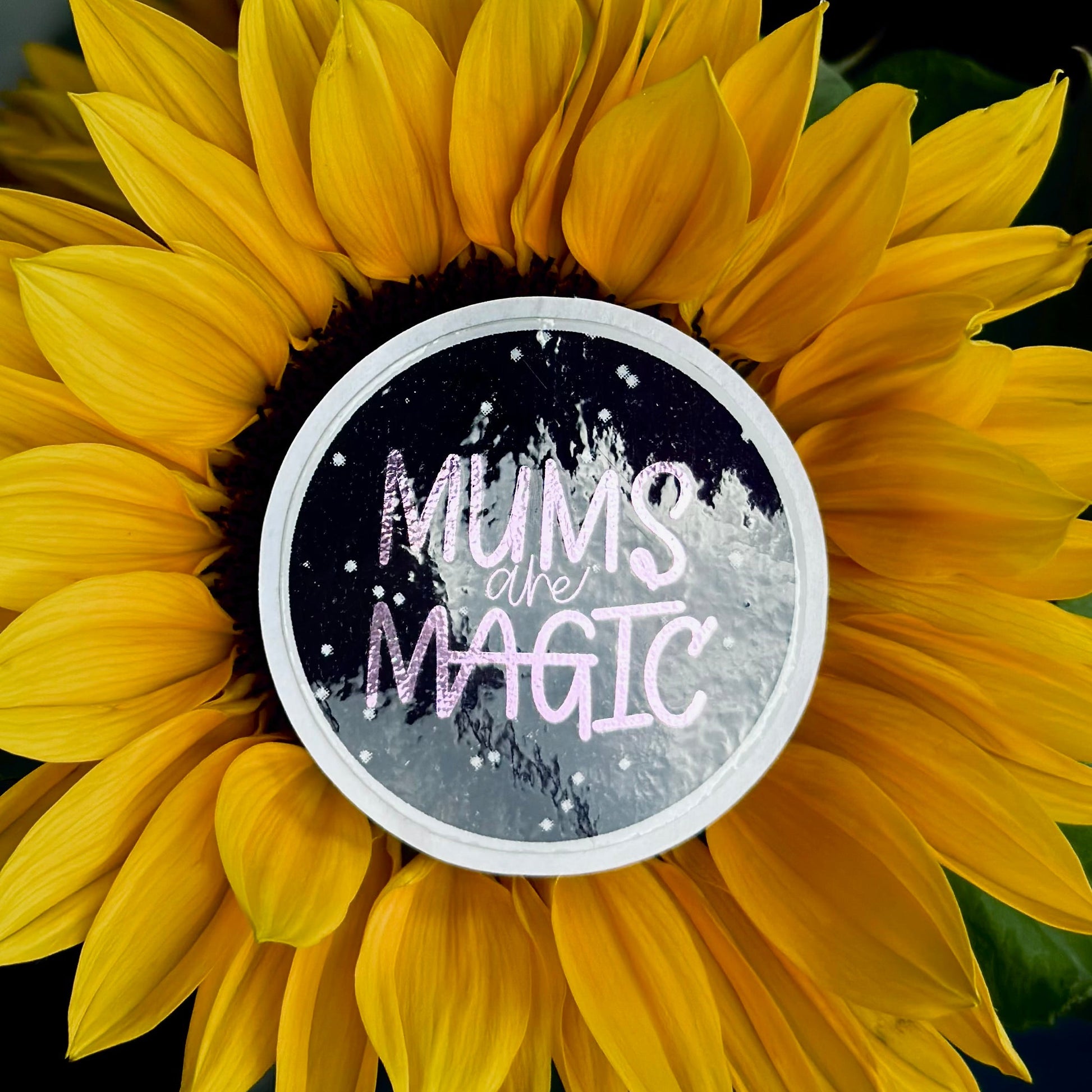 A round sticker placed on the center of a large yellow sunflower. The sticker reads “MUMS are MAGIC” in pink and white lettering over a dark starry background.