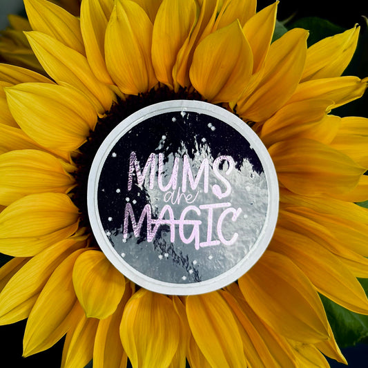 A round sticker placed on the center of a large yellow sunflower. The sticker reads “MUMS are MAGIC” in pink and white lettering over a dark starry background.