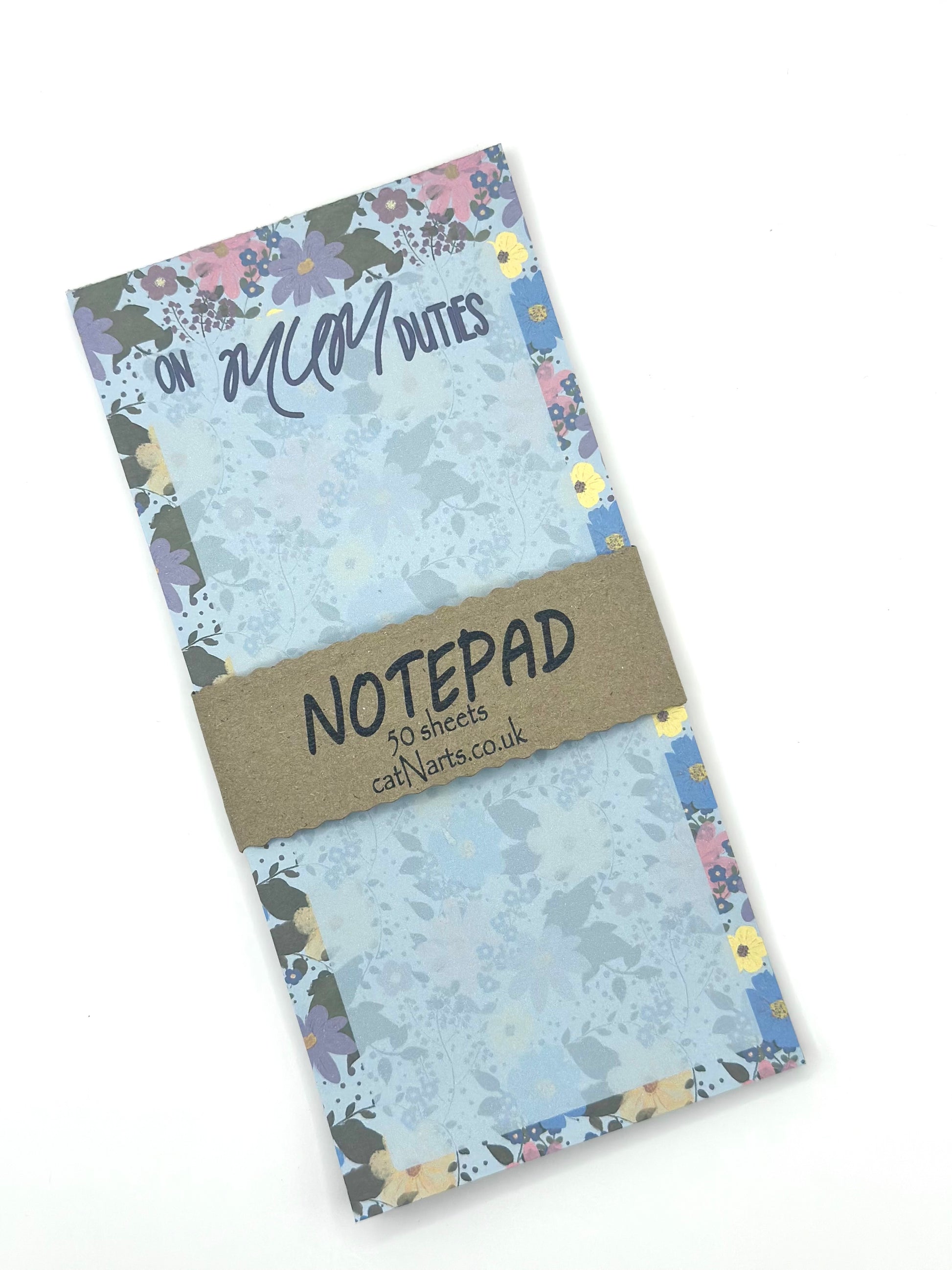 A floral-patterned notepad with a text 'on mum duties' with a brown paper band reading 'NOTEPAD' and '50 sheets 