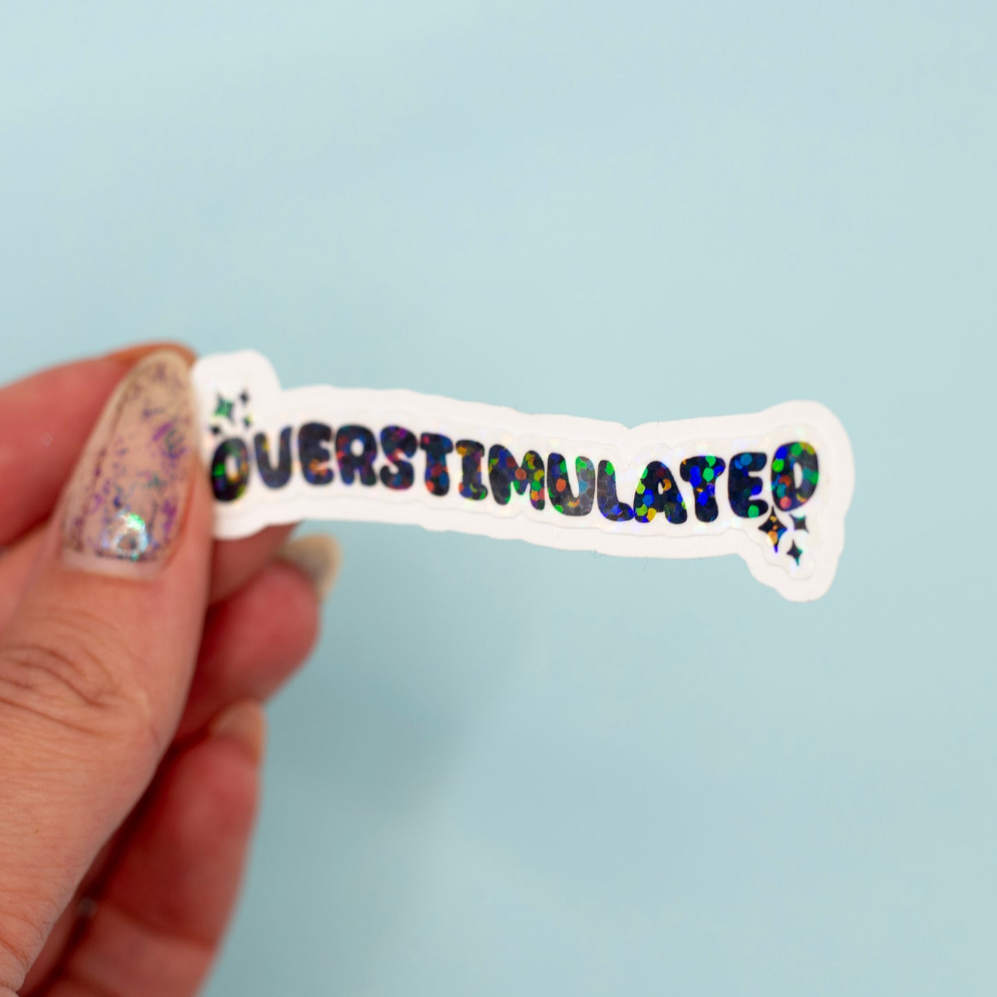  a die-cut sticker that reads 'OVERSTIMULATED' in colorful holographic lettering on a white background, set against a solid light blue backdrop.