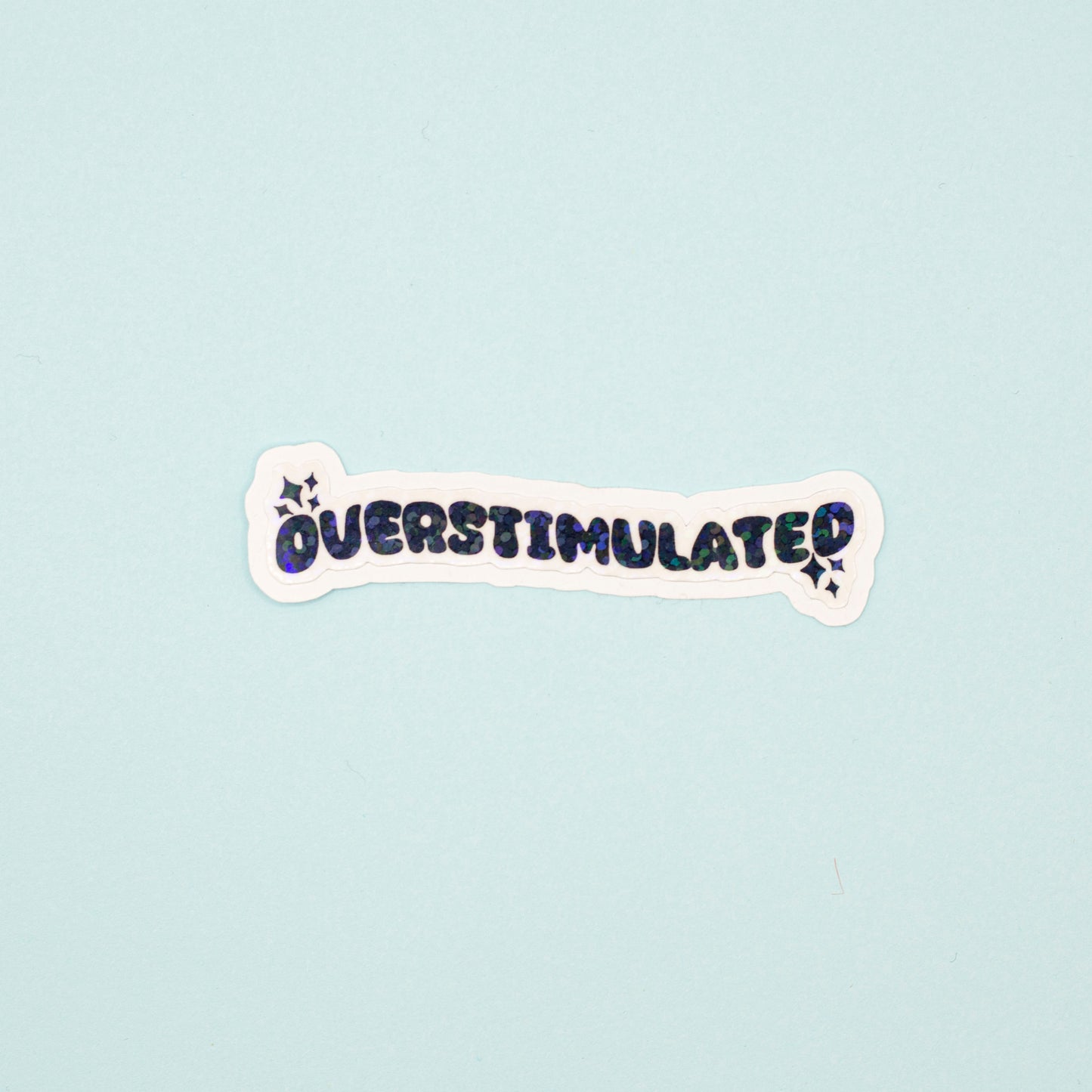 A die-cut sticker with the word 'OVERSTIMULATED' in black bubble letters and a holographic dot overlay on a white background,  shown on a light blue background.